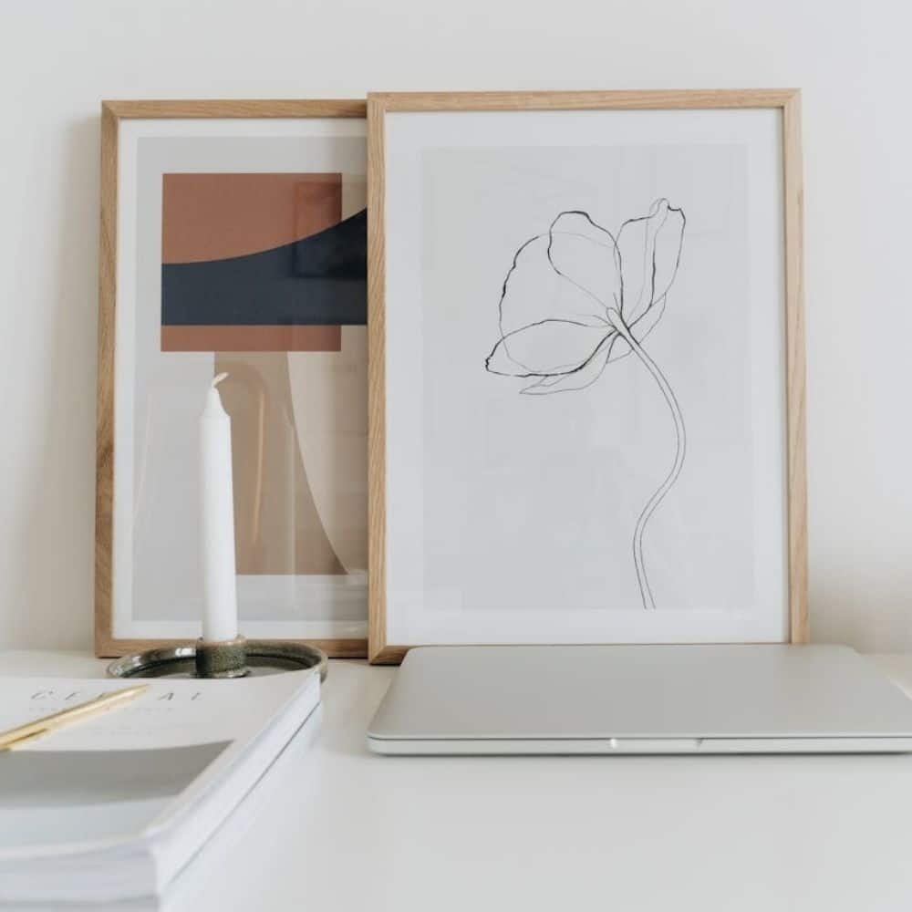 Two framed abstract art pieces lean against a wall on a white desk with a laptop and notebooks