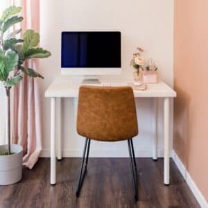 A minimalist home office featuring a white desk, a black computer monitor, a brown chair, a potted plant, and pink curtains.