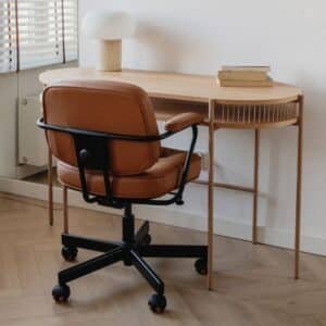A brown leather office chair with black accents sits in front of a light wood desk with a book and a cup on it.