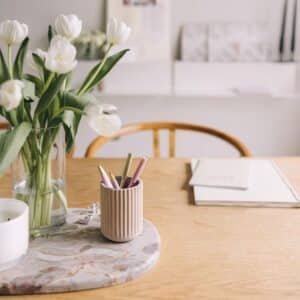 A vase filled with white tulips is placed on a table beside a small hexagonal holder containing pens and a notebook, with shelves and a chair blurred in the background.