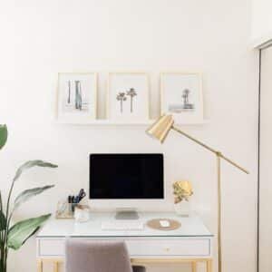 A minimalist home office setup with a white desk, computer monitor, and a gold desk lamp, all against a white wall with three framed prints on a shelf above.